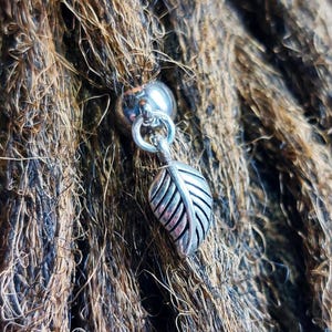 May include: Silver dreadlock bead with a leaf charm. The bead is round and has a small hole in the center. The leaf charm is attached to the bead with a small loop.