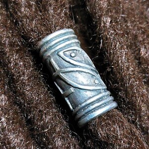 May include: A silver-colored dreadlock bead with an etched design. The cylindrical bead is adorned with geometric patterns and is set against a backdrop of brown dreadlocks. The bead adds a decorative element to the hair.