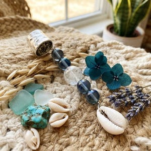 May include: A decorative hanging ornament featuring a hammered silver-toned metal cap, a strand of translucent blue and clear crackle glass beads, and a white cowrie shell. The piece is accented with dried teal hydrangea petals and lavender.