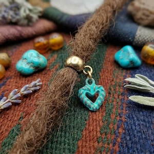 May include: A brown dreadlock with a gold bead and a turquoise heart charm. The charm has a distressed finish. Turquoise stones and amber beads are scattered around the dreadlock on a patterned fabric.