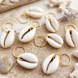 May include: A collection of eight off-white cowrie shells, each attached to a small, circular gold-toned metal ring. The shells feature a smooth, polished surface with a characteristic central slit, and the metal rings are thin and polished.