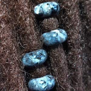 Op de afbeelding: Vier blauwgrijze edelsteen kralen met een gevlekt patroon zijn geregen op bruin dreadlocks.