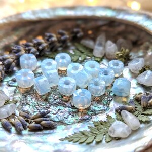 May include: A collection of small, translucent, pale blue faceted beads with a central hole, arranged in a cluster. The beads have a smooth, polished surface and a rounded, multi-faceted shape that reflects light, creating a soft glow.