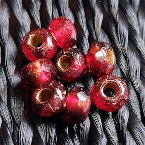 May include: Eight red glass beads with gold flecks and a hole in the center. The beads are on a black braided surface.