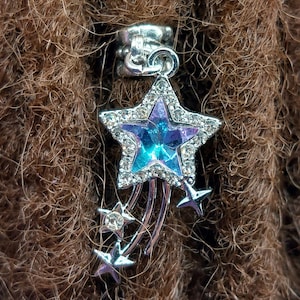 May include: A silver star charm with a blue and purple crystal in the center. The star is surrounded by smaller silver stars and is attached to a silver bead.