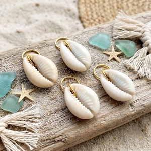 May include: Four white cowrie shell charms, each featuring a gold-toned metal jump ring attached to the top. The shells have a smooth, glossy finish with a central slit and ribbed texture, arranged on a weathered piece of driftwood.