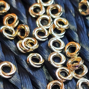 May include: A close-up of a group of gold-colored metal hair rings. The rings are arranged in a random pattern on a black background.
