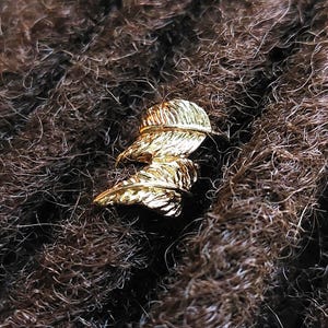 May include: A close-up shot showcases a gold-colored hair accessory, shaped like two overlapping feathers. The accessory is set against a backdrop of dark brown hair, creating a contrast that highlights the intricate details of the metalwork.