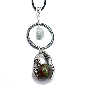 May include: A silver pendant necklace with a green and red stone held in a silver claw design. The necklace has a black cord and a silver circle with a clear stone hanging above the claw.