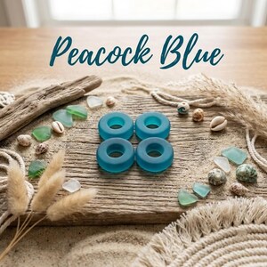 May include: Four circular, peacock blue glass rings arranged in a square formation. The text "Peacock Blue" is centered above the rings. The items are displayed on a weathered wooden surface with scattered seashells, sea glass, and dried grasses.