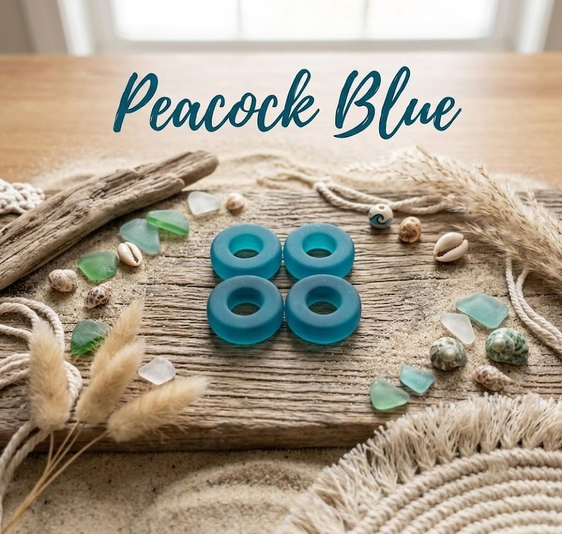 May include: Four circular, peacock blue glass rings arranged in a square formation. The text "Peacock Blue" is centered above the rings. The items are displayed on a weathered wooden surface with scattered seashells, sea glass, and dried grasses.