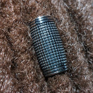 May include: A silver thimble with a textured surface of small, evenly spaced bumps. The thimble is cylindrical with a slightly flared top and bottom, resting on a brown, textured surface.
