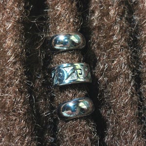 May include: Three silver dreadlock beads, one plain, one with a decorative design, and one plain. The beads are on brown dreadlocks.