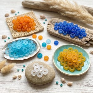 May include: A collection of small, round, translucent ring-shaped beads in orange, blue, light blue, yellow, and white. The beads are arranged in piles on a woven square mat, a piece of driftwood, a shell, a ceramic dish, and a smooth stone.