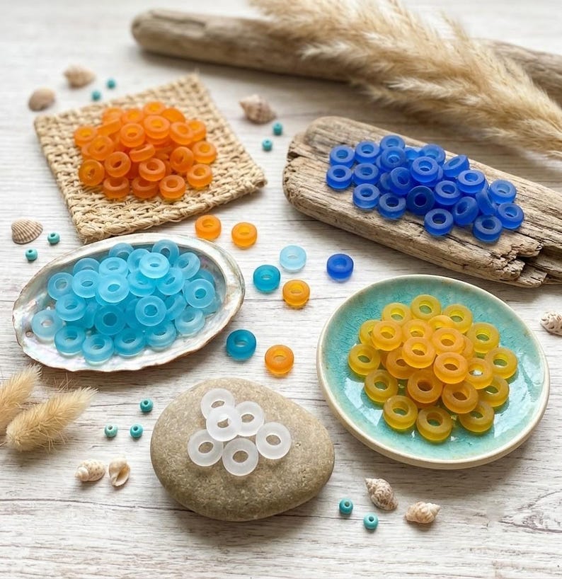 May include: A collection of small, round, translucent ring-shaped beads in orange, blue, light blue, yellow, and white. The beads are arranged in piles on a woven square mat, a piece of driftwood, a shell, a ceramic dish, and a smooth stone.
