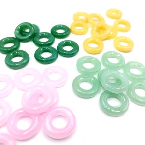 May include: A collection of colorful, ring-shaped beads in shades of green, yellow, pink, and light green. The beads are smooth and appear to be made of a polished stone or glass material. The beads are scattered on a white surface.