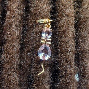 May include: A gold-tone hair tie with a pink crystal cat charm. The cat charm has a bow and a tail.
