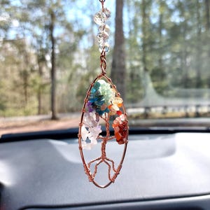 May include: A copper wire tree of life car charm with colorful gemstones. The charm features a tree design with a variety of stones in shades of blue, green, purple, orange, and red. Clear crystal beads and a black cord complete the design.