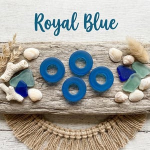 May include: Three royal blue circular glass beads with center holes, surrounded by various white seashells, pieces of light green and dark blue sea glass, and dried coral fragments arranged on a textured wooden surface.