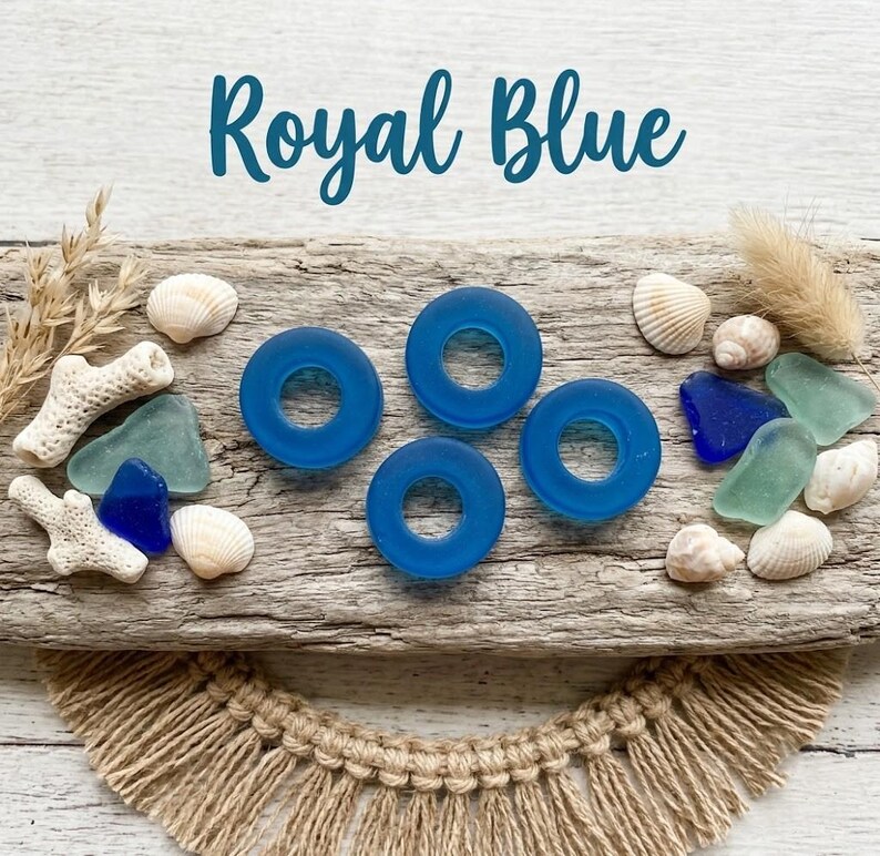 May include: Three royal blue circular glass beads with center holes, surrounded by various white seashells, pieces of light green and dark blue sea glass, and dried coral fragments arranged on a textured wooden surface.