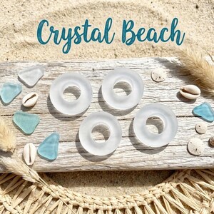 May include: Four frosted white glass rings arranged on a weathered wooden board, accompanied by scattered light blue sea glass pieces and small white seashells. The text Crystal Beach is displayed in a dark teal script font above the items.