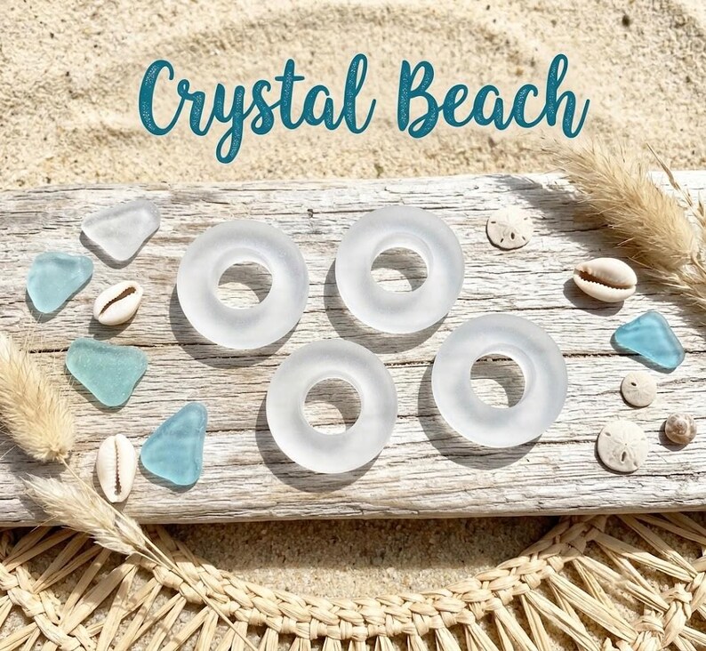 May include: Four frosted white glass rings arranged on a weathered wooden board, accompanied by scattered light blue sea glass pieces and small white seashells. The text Crystal Beach is displayed in a dark teal script font above the items.