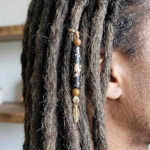 May include: A gold-colored dreadlock bead with a black and gold design, featuring a feather charm and wooden beads. The accessory is attached to brown dreadlocks.