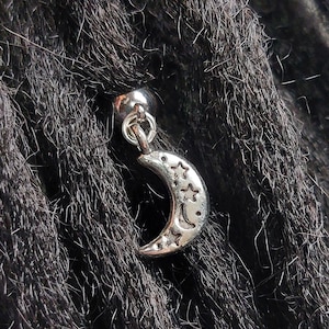 May include: Silver crescent moon charm with stars hanging from a silver bead. The charm is attached to a dreadlock.