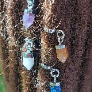 May include: Four silver dreadlock beads with different colored crystal charms. The charms are in the shape of a point and are amethyst, orange, white, and blue.