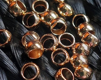 May include: A collection of gold-colored metal beads, designed for use in hair braiding. The beads are round with a hole in the center, and they are arranged on a black background.