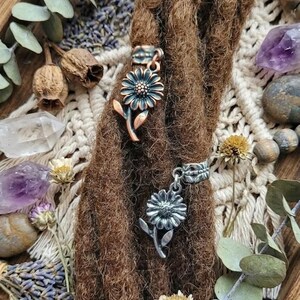May include: Close-up of brown dreadlocks adorned with two daisy-shaped hair accessories. One is copper-toned, the other silver. The accessories are set against a backdrop of dried flowers, crystals, and a wooden tray, creating a bohemian aesthetic.