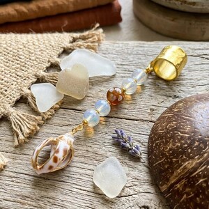 May include: A handcrafted gold-toned ring with a seashell charm, glass beads, and a gold-toned ring. The ring is adorned with a seashell, opalescent beads, and a decorative gold-toned ring. The background includes sea glass and a sprig of lavender.