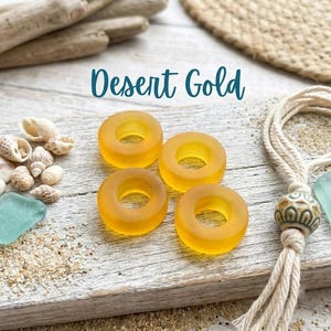 May include: Four translucent, circular, golden-yellow glass beads with a smooth, matte finish. The beads are arranged in a square formation. The text "Desert Gold" is printed in a dark teal script font above the beads.