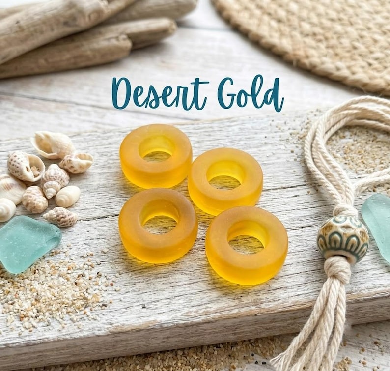 May include: Four translucent, circular, golden-yellow glass beads with a smooth, matte finish. The beads are arranged in a square formation. The text "Desert Gold" is printed in a dark teal script font above the beads.