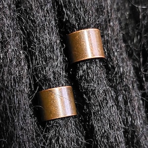 May include: Close-up of two copper-colored hair cuffs on dark, textured hair. The cylindrical cuffs are smooth and metallic, contrasting with the rough, black strands. The image highlights the hair accessories and their placement.