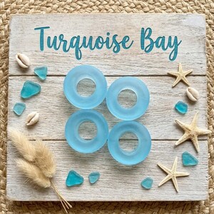 May include: A square wooden sign with the text Turquoise Bay in teal script. Four light blue circular glass rings are arranged in a square pattern in the center. The sign is decorated with small white seashells, three beige starfish, and several small teal sea glass pieces, along with a cluster of dried tan grass stems.