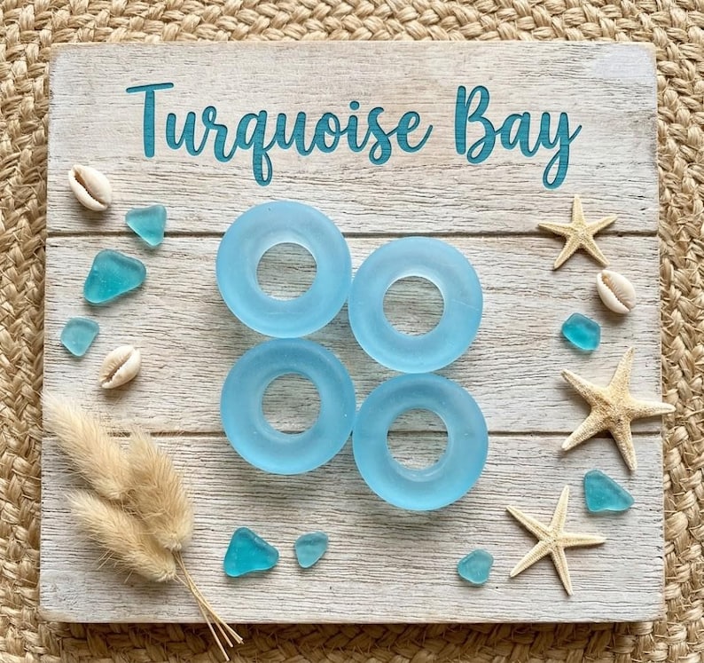 May include: A square wooden sign with the text Turquoise Bay in teal script. Four light blue circular glass rings are arranged in a square pattern in the center. The sign is decorated with small white seashells, three beige starfish, and several small teal sea glass pieces, along with a cluster of dried tan grass stems.