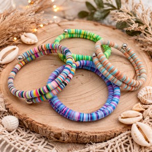May include: A collection of colorful, beaded bracelets with a variety of striped patterns. The bracelets are made of small, flat beads in shades of pink, blue, green, and orange. Each bracelet has a silver-colored metal clasp. The bracelets are displayed on a wooden surface with seashells.