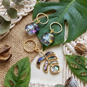 May include: Gold-toned keychains and a pendant featuring iridescent, heart-shaped glass beads and a leaf design. The keychains have a simple ring attachment. The leaf pendant has a gold outline with iridescent abalone shell inlays. Green leaves and natural elements surround the jewelry.