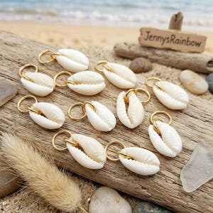 May include: A collection of white seashell charms with gold-colored hoops, arranged on a piece of driftwood. The charms are oval-shaped and have a natural, textured appearance. The background includes sand and a small wooden sign.