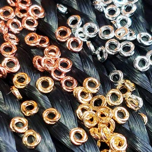 May include: A close-up of a variety of small, round, metal rings in gold, silver, and rose gold. The rings are scattered on a black surface.