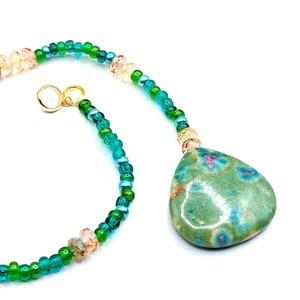 May include: A handcrafted necklace featuring a large, teardrop-shaped pendant in shades of green, blue, and pink. The pendant is suspended from a beaded chain with green, teal, and peach-colored beads. A gold-colored clasp secures the necklace.