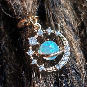 May include: A gold-colored pendant featuring a celestial design. The pendant has a central blue orb, encircled by a gold band, and surrounded by a crescent shape adorned with clear, sparkling crystals. The pendant is suspended from a gold chain.