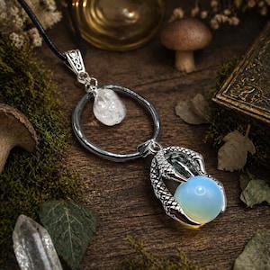 May include: A silver-toned pendant necklace featuring a clear quartz crystal and a claw holding a blue-green orb. The pendant hangs from a black cord. The background includes moss, leaves, and a wooden surface.