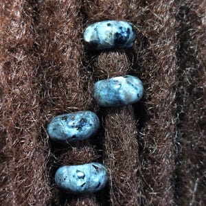Op de afbeelding: Vier blauwgrijze edelsteen kralen met donkere vlekken zijn geregen op bruin dreadlocks.