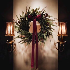 May include: A festive Christmas wreath made of green pine needles and pinecones, adorned with a burgundy ribbon. Two gold sconces with lit lamps flank the wreath, creating a warm, inviting ambiance.
