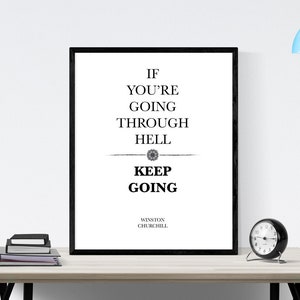 May include: Black and white motivational quote print featuring the text "If you're going through hell, keep going" attributed to Winston Churchill. The print is framed in a black frame and is displayed on a wooden table with a black clock and a stack of books.