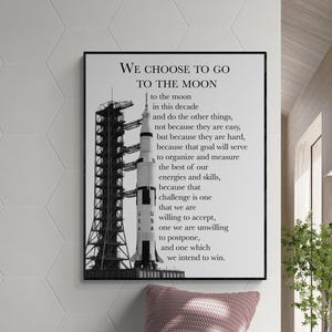 May include: A framed black and white print featuring a rocket and the text "WE CHOOSE TO GO TO THE MOON". The print is displayed above a wooden bench with a pink pillow and blanket. The room has a modern design.