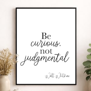 May include: A framed print with the quote "Be curious, not judgmental" in black script on a white background. The print is in a black frame and includes the name "Walt Whitman" below the quote.