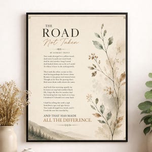 May include: A framed wall art print featuring the poem The Road Not Taken by Robert Frost. The text is printed in black on a cream background, accompanied by a watercolour illustration of a delicate leafy branch and a landscape of pine trees.